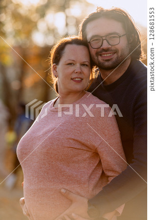 A man lovingly embraces his pregnant wife in the park, sharing a peaceful moment of warmth and affection A man lovingly embraces his pregnant wife in the park, sharing a peaceful moment of warmth and affection 128681553