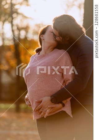 A man lovingly embraces his pregnant wife in the park, sharing a peaceful moment of warmth and affection A man lovingly embraces his pregnant wife in the park, sharing a peaceful moment of warmth and affection 128681558