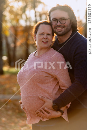 A man lovingly embraces his pregnant wife in the park, sharing a peaceful moment of warmth and affection A man lovingly embraces his pregnant wife in the park, sharing a peaceful moment of warmth and affection 128681559