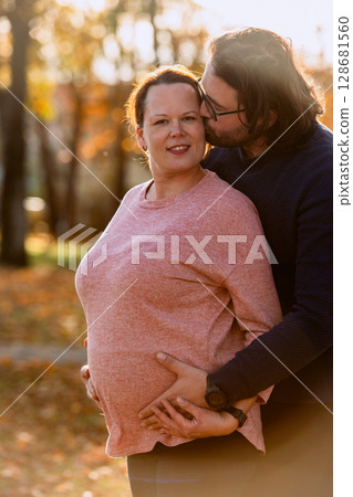 A man lovingly embraces his pregnant wife in the park, sharing a peaceful moment of warmth and affection  128681560