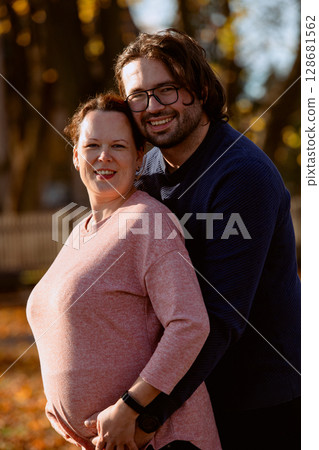 A man lovingly embraces his pregnant wife in the park, sharing a peaceful moment of warmth and affection  128681562