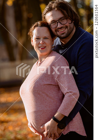A man lovingly embraces his pregnant wife in the park, sharing a peaceful moment of warmth and affection A man lovingly embraces his pregnant wife in the park, sharing a peaceful moment of warmth and affection 128681564
