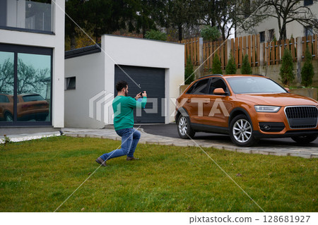A man meticulously washes and cleans his car in the driveway of his home, showcasing pride in his vehicle and outdoor space. 128681927