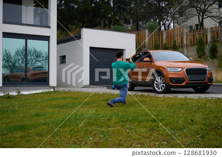 A man meticulously washes and cleans his car in the driveway of his home, showcasing pride in his vehicle and outdoor space. 128681930