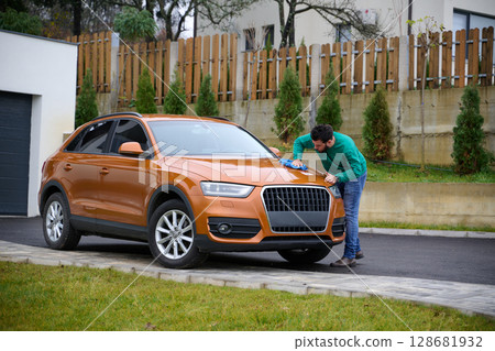 A man meticulously washes and cleans his car in the driveway of his home, showcasing pride in his vehicle and outdoor space. 128681932