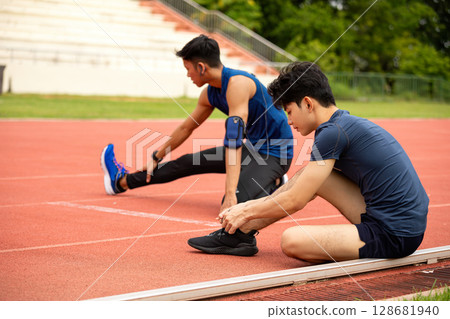 Two male athletes sitting on red running track tying shoelaces and stretching legs before workout Two male athletes sitting on red running track tying shoelaces and stretching legs before workout 128681940