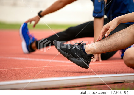 Close-up of two male athletes stretching legs on red running track focusing on sneakers and posture 128681941