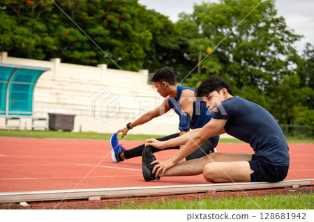 Two young male athletes stretching on red running track with focused expressions and warm-up posture Two young male athletes stretching on red running track with focused expressions and warm-up posture 128681942