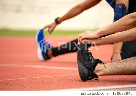 Close-up of two male athletes stretching legs on red track with focus on black running shoe and hand 128681943