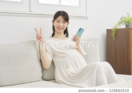 Pregnant woman with smartphone, peace sign, living room 128682010