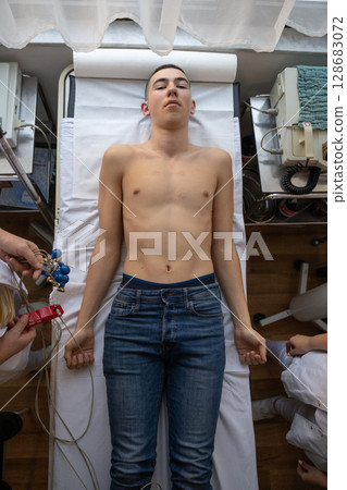 A female doctor performs an ECG on a young man in a hospital room, carefully monitoring his heart health during a routine checkup. A female doctor performs an ECG on a young man in a hospital room, carefully monitoring his heart health during a routine checkup. 128683072