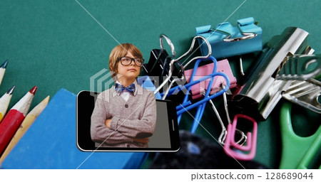 Image of smart caucasian schoolboy in smartphone moving over school stationery on desk Image of smart caucasian schoolboy in smartphone moving over school stationery on desk 128689044