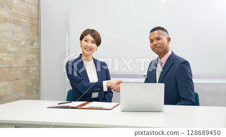 A black male businessman and a Japanese female staff member shaking hands in front of a whiteboard 128689450