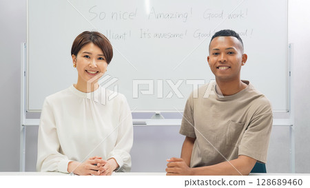 A black man and a Japanese woman standing side by side looking at the camera in a classroom 128689460