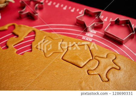 Rolled-out gingerbread dough with metal cookie cutters in shape of stars, houses, and gingerbread men on red mat Rolled-out gingerbread dough with metal cookie cutters in shape of stars, houses, and gingerbread men on red mat 128690331
