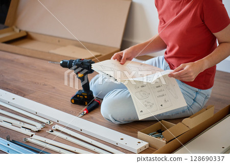 Woman sitting on floor reading instruction manual during furniture assembling. Person sitting on floor near tools and parts of new bed. Concept of DIY home improvement and furniture installation 128690337