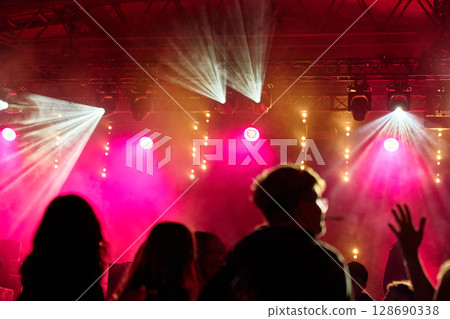 Crowd enjoying concert with bright lights and smoke on stage. Silhouettes of people dancing at night in front of concert stage with spotlights. Concept of live music event and entertainment 128690338