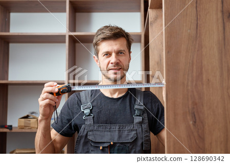 Man in work overalls holding measuring tape during assembling wooden wardrobe. Concept of DIY home improvement, handyman work and self furniture installation process 128690342