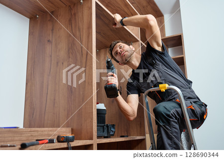 Man wearing work overalls assembling wooden wardrobe with drill and tools. Carpenter standing on step ladder, furniture installation process 128690344