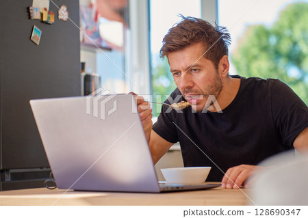 Man has breakfast while working on laptop at home kitchen. Person eating porridge. Work and life balance and busy morning concept 128690347