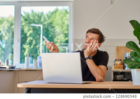 Man working on laptop at home and using remote control to adjust temperature on air conditioner. Person struggling with heat while working at home office 128690348