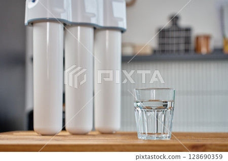 Modern water filtration system with three cartridges and glass of clean water on kitchen table. Home water purification, healthy lifestyle and drinking water safety concept 128690359