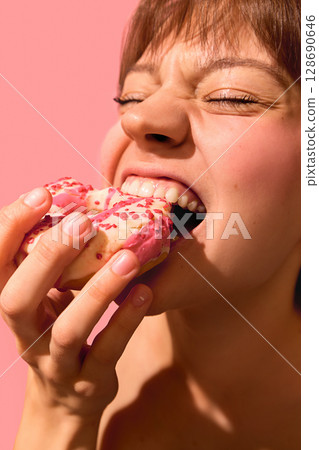 Close-up of woman biting frosted pink donut with emotional joyful intensity Close-up of woman biting frosted pink donut with emotional joyful intensity 128690646