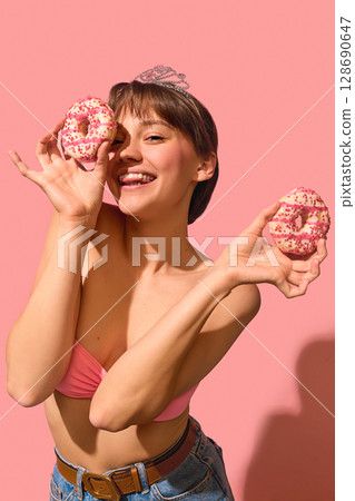 Smiling woman in tiara holding donuts near eyes with playful joyful expression 128690647