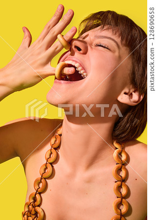 Joyful woman biting bagel rings from fingers in surreal yellow snack fashion 128690658