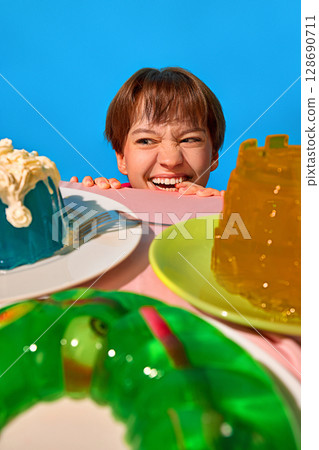 Excited woman peeking over table with colorful jelly desserts in playful composition Excited woman peeking over table with colorful jelly desserts in playful composition 128690711