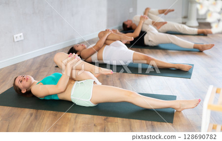 Man and woman doing yoga with group in studio, performing hamstring and low back stretching Ardha Pawanmuktasana 128690782