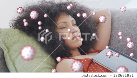 Lying woman in orange polka-dot dress holding book on sofa at home, with floating virus overlay 128691981