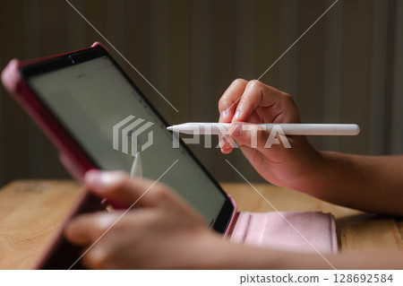 Cute Asian girl drawing using electronic tablet and stylus pen. Asian elementary school girl using digital tablet sitting in living room at home. Cute Asian girl drawing using electronic tablet and stylus pen. Asian elementary school girl using digital tablet sitting in living room at home. 128692584