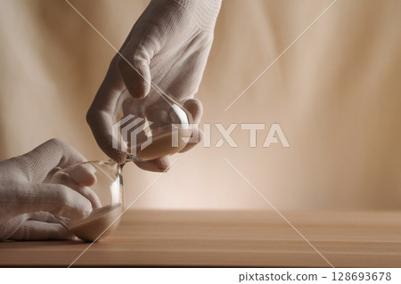 A glove-covered hand holding a tipped hourglass on a wooden surface, symbolizing the flow of time, creativity, and patience in a contemporary minimalist style. 128693678
