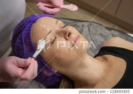 Peeling procedure in beauty salon. Lying woman while rejuvenation treatment. Collagen therapy in spa salon. Woman face while applying exfoliation by brush. Cosmetology clinic. Anti aging procedure. Peeling procedure in beauty salon. Lying woman while rejuvenation treatment. Collagen therapy in spa salon. Woman face while applying exfoliation by brush. Cosmetology clinic. Anti aging procedure. 128693805
