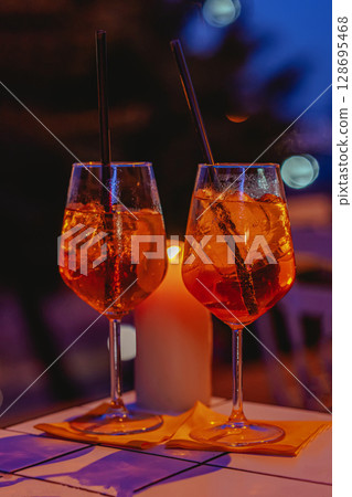 Two vibrant aperol spritz cocktails with ice and straws on a table by candlelight, glowing in warm evening tones. Concept of aperol spritz as romantic drink for summer nights 128695468