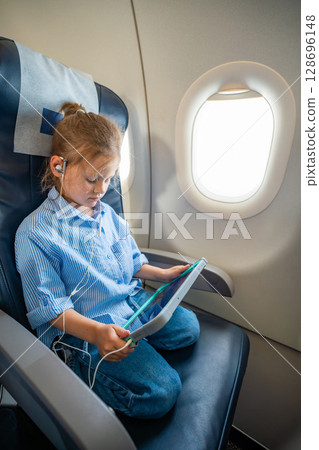 Little girl watching a cartoon on a tablet with headphones during an airplane flight. Digital entertainment, child focus, and quiet family travel experience. 128696148