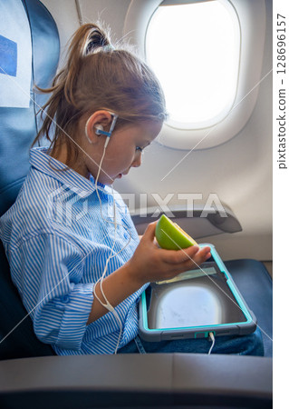Little girl watching cartoons on tablet and eating green apple during airplane flight. Candid moment of child entertainment and healthy snack onboard. Little girl watching cartoons on tablet and eating green apple during airplane flight. Candid moment of child entertainment and healthy snack onboard. 128696157