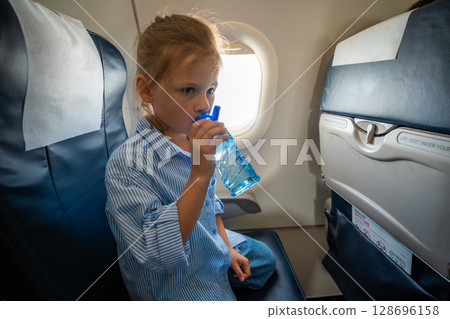 Little girl drinking water from a plastic bottle during a flight. In-flight hydration, travel routine, and caring for body needs while flying Little girl drinking water from a plastic bottle during a flight. In-flight hydration, travel routine, and caring for body needs while flying 128696158