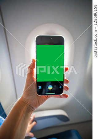 Close-up of a hand holding a smartphone with green screen camera active, recording view from airplane window. Travel content creation, mobile photography, and mockup preparation in flight Close-up of a hand holding a smartphone with green screen camera active, recording view from airplane window. Travel content creation, mobile photography, and mockup preparation in flight 128696159