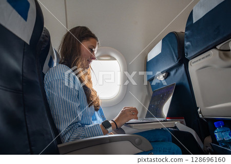 Young woman working on a laptop during a flight, seated by the airplane window. Remote work, travel productivity, and digital lifestyle on the go Young woman working on a laptop during a flight, seated by the airplane window. Remote work, travel productivity, and digital lifestyle on the go 128696160
