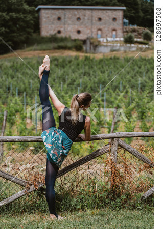 Woman Practicing Leg Stretch by a Vineyard Fence 128697508