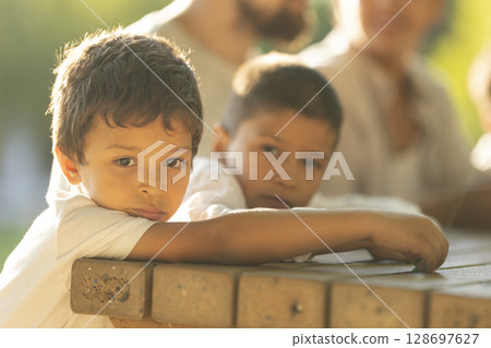 Pensive latin american brothers leaning on picnic table with family in background 128697627