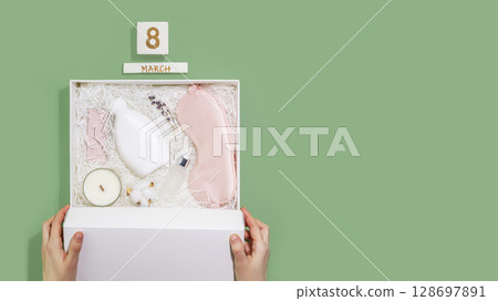 Womans day holiday flat lay with female hands hold white gift box with sleep care set. Self care package, craft gift box with mask for sleep, aroma candle and bubble bath. Flat lay, Copy space 128697891