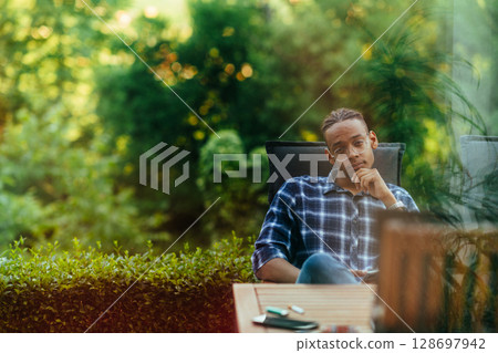 An African American man sitting in front of the house on a modern terrace and stares thoughtfully at the camera.  128697942