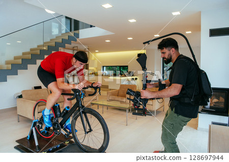 A cameraman filming an athlete riding a triathlon bike on a simulation machine in a modern living room. Training in pandemic conditions 128697944