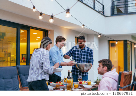 A group of friends and family barbecue together in the evening on the terrace in front of a large modern house. A group of friends and family barbecue together in the evening on the terrace in front of a large modern house. 128697951