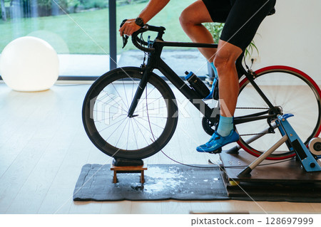 A triathlete riding a triathlon bike on a machine simulation in a modern living room. Training during pandemic conditions. 128697999
