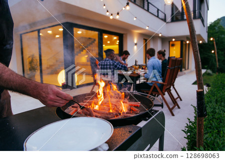 A group of friends and family barbecue together in the evening on the terrace in front of a large modern house. A group of friends and family barbecue together in the evening on the terrace in front of a large modern house. 128698063