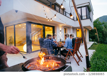 A group of friends and family barbecue together in the evening on the terrace in front of a large modern house.  128698065